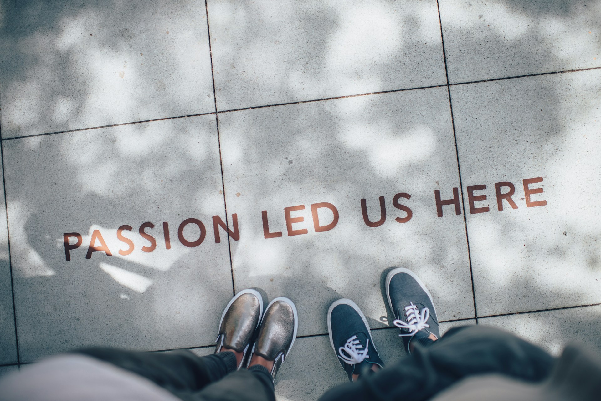 Two pairs of shoes standing near pavement text that reads “Passion led us here,” with dappled sunlight creating shadow patterns.